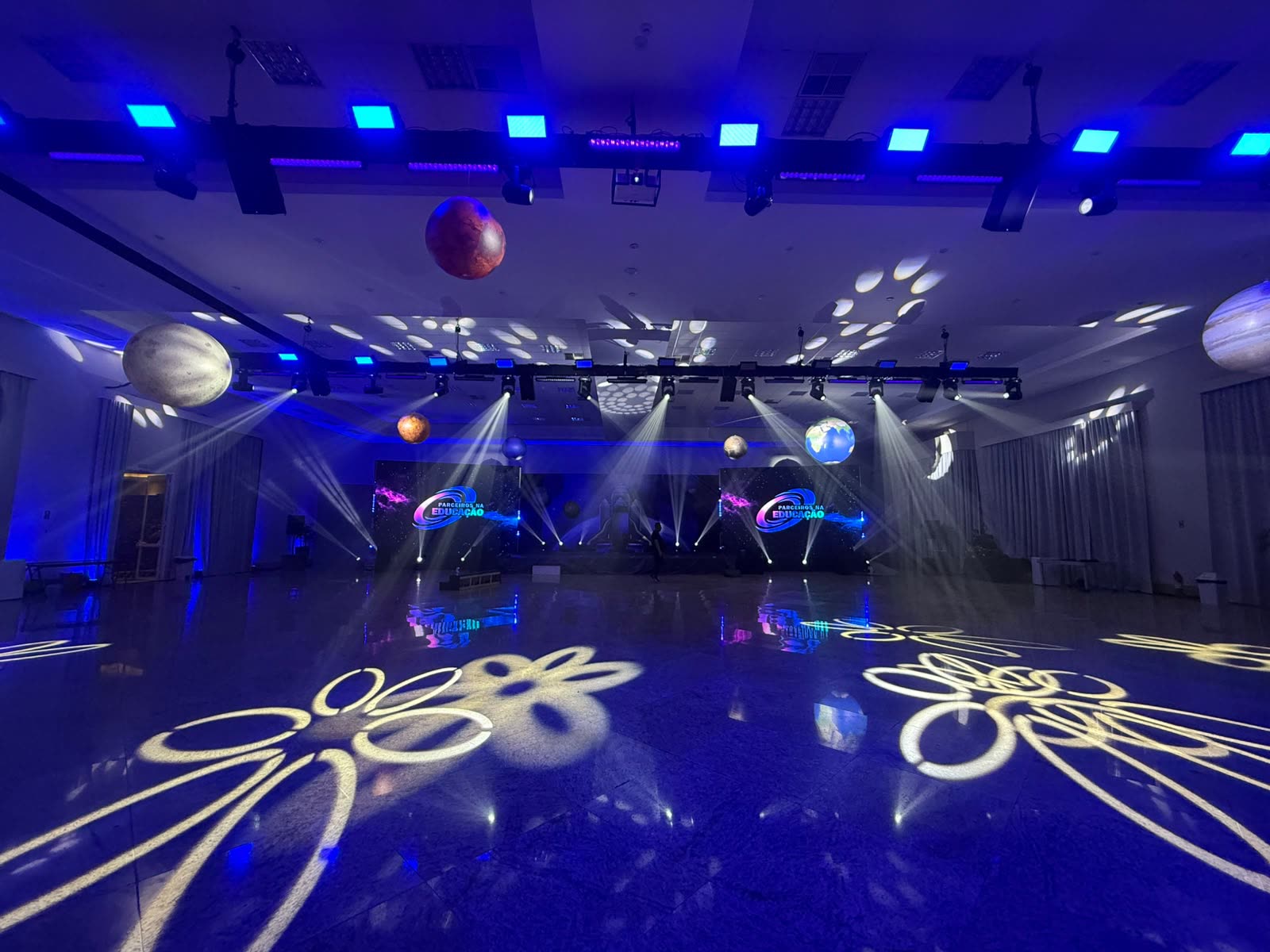 Salão montado com moving heads, gobos no piso e planetas cenográficos suspensos